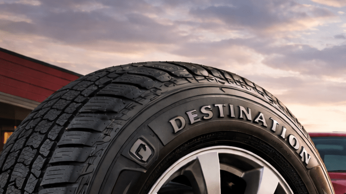 Primer plano de neumático Firestone frente a taller automotriz al atardecer, con camioneta y SUV sobre asfalto mojado iluminado por luz cálida.