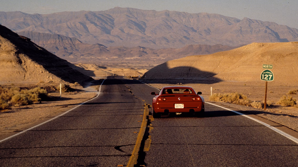 Ferrari F355 rojo en carretera desértica, deportivo clásico italiano fotografiado desde atrás en un paisaje montañoso