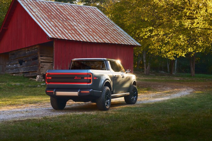 Scout Terra pick-up eléctrica gris con granero rojo de fondo, vista trasera con luces LED encendidas.