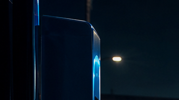 Auto eléctrico cargándose de noche con cable de luz azul e impuestos al fondo