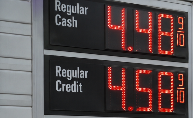 Mujer cargando combustible en gasolinera mientras avión comercial sobrevuela con precio de gasolina a $4.48