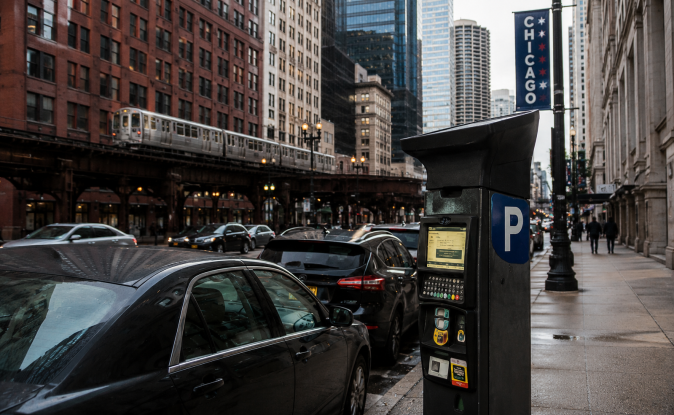 Parquímetro moderno en calle del centro de Chicago, autos estacionados junto a vía elevada del tren y rascacielos de fondo.