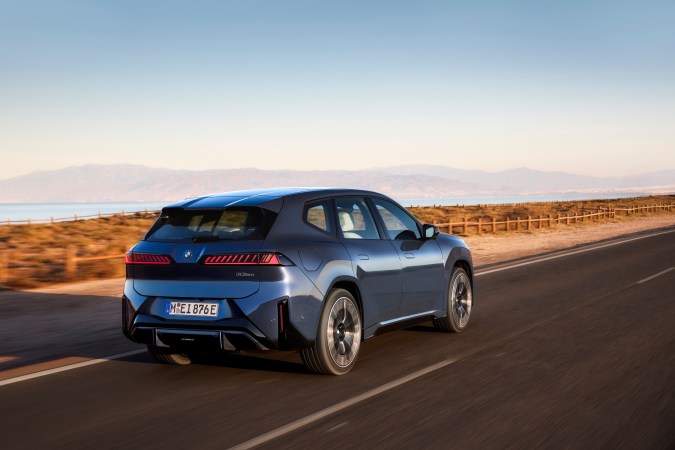 BMW iX3 50 xDrive azul circulando por carretera costera, SUV eléctrico con diseño moderno, vista trasera y luces LED.