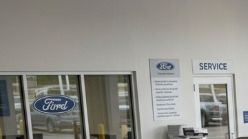 Fotografía de plano medio que muestra el interior de un concurrido taller de servicio de concesionario Ford, capturando la actividad de los técnicos trabajando en varios vehículos de la marca, como el Mustang Mach-E y el Ford Bronco Sport