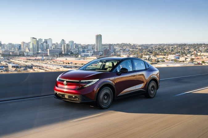 Crossover eléctrico rojo tipo Nissan Leaf 2026 circulando en autopista elevada con panorámica de ciudad moderna al fondo