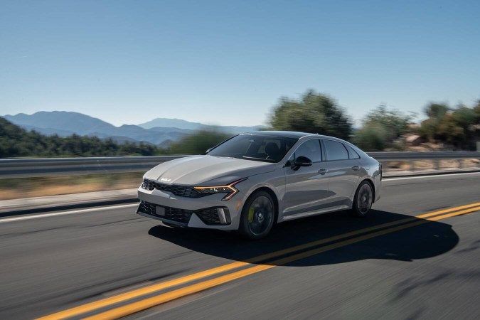 Kia K5 2026 plateado en movimiento en autopista. Sedán elegante con diseño deportivo y paisaje montañoso