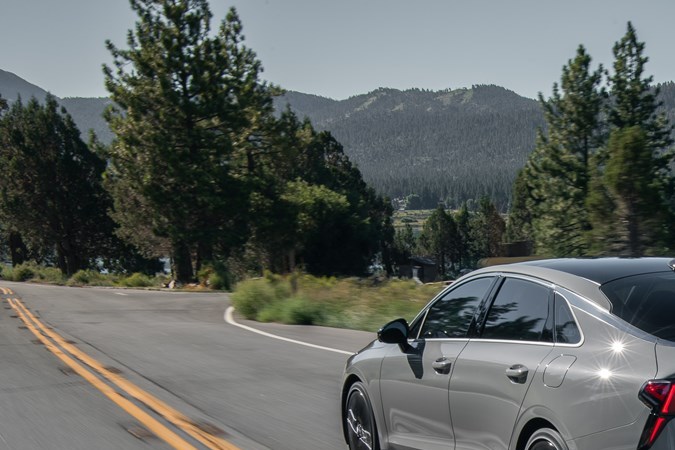 Vista trasera Kia K5 2026 en carretera con paisaje montañoso y lago.