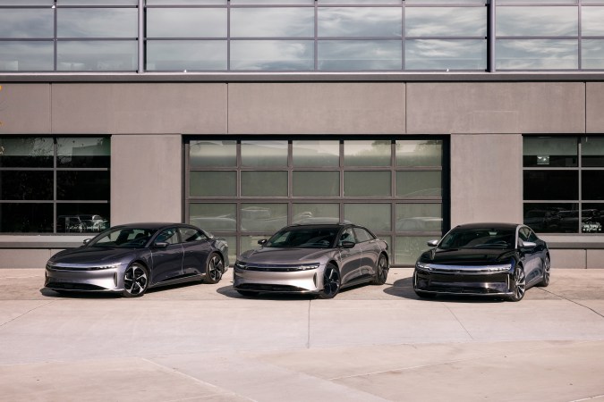 Tres sedanes Lucid Air eléctricos en gris, plata y negro estacionados frente a edificio moderno de la marca.