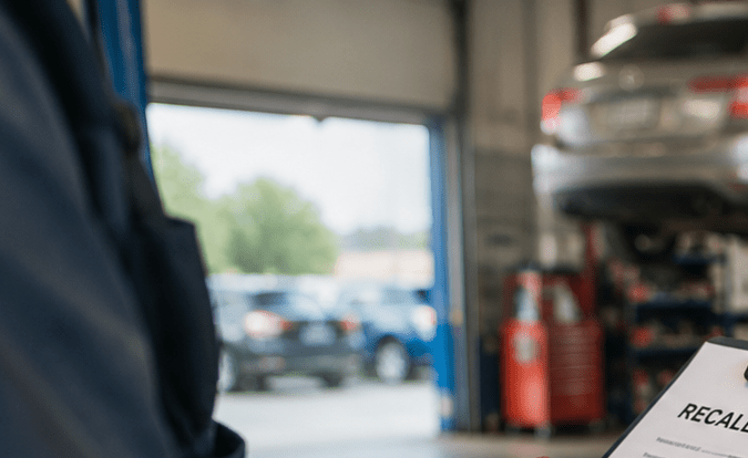 Mecánico sosteniendo aviso de recall de vehículo en taller automotriz, con capó abierto y coche en elevador.