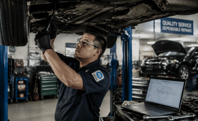 Técnico automotriz certificado ASE inspecciona parte inferior de vehículo elevado con laptop de diagnóstico en taller