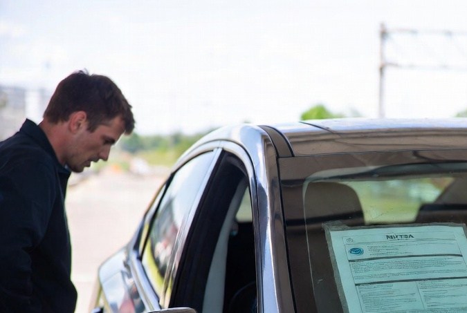 Hombre revisando documento de recall pegado en el parabrisas de un coche en estación de servicio.