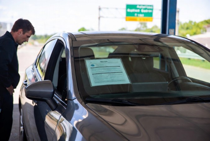 Hombre revisando documento de recall pegado en el parabrisas de un coche en estación de servicio.