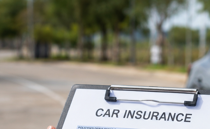 Formulario de seguro de coche en portapapeles con auto gris de fondo. Contratación de póliza de seguro automovilístico