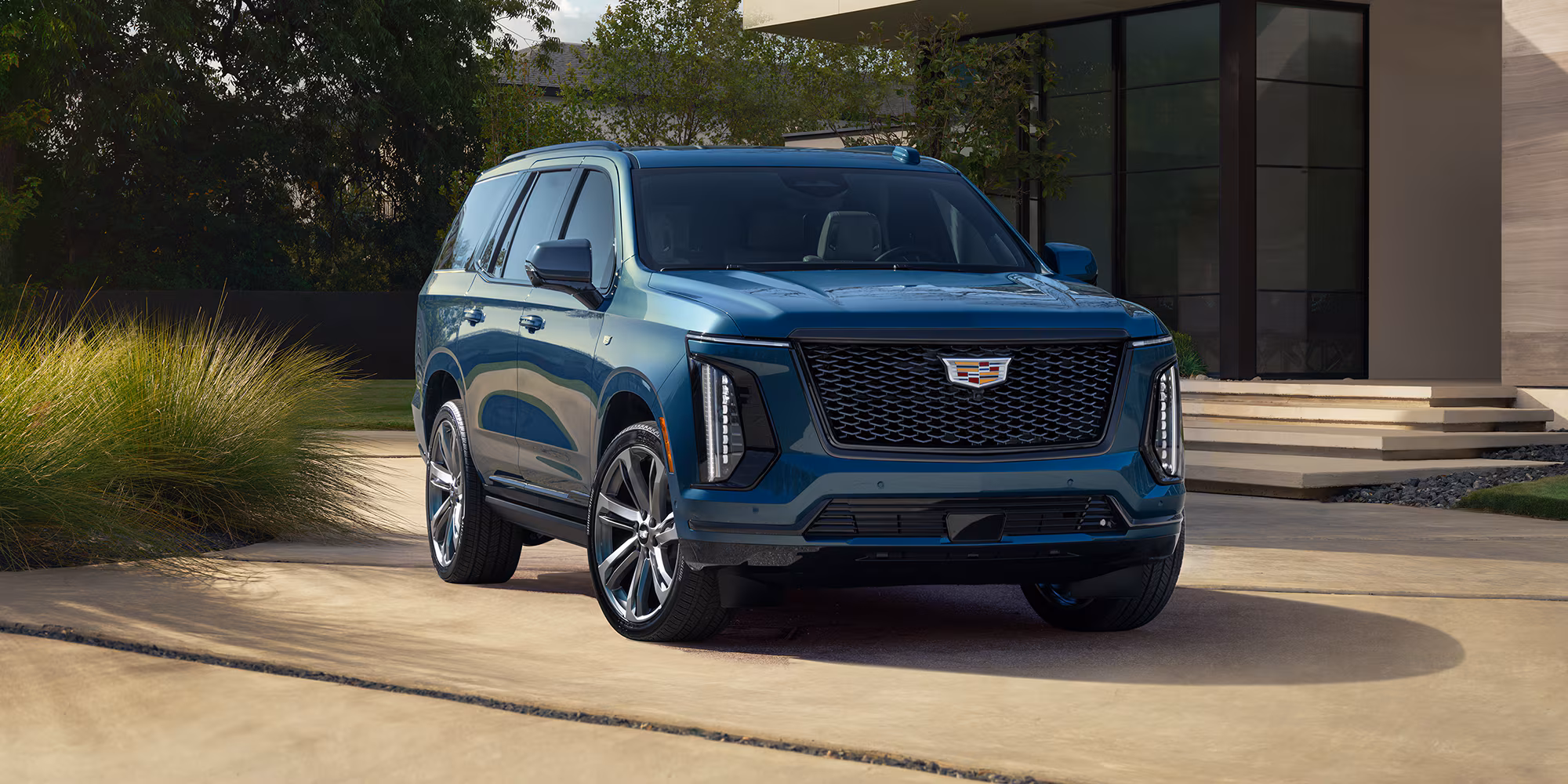 Vista delantera del Cadillac Escalade 2026 azul, SUV de lujo grande estacionado en entrada de casa moderna con diseño elegante.