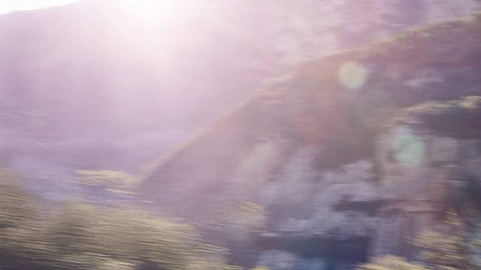 McLaren 570S gris circulando a gran velocidad por carretera de montaña con curvas, vista trasera dinámica al atardecer.