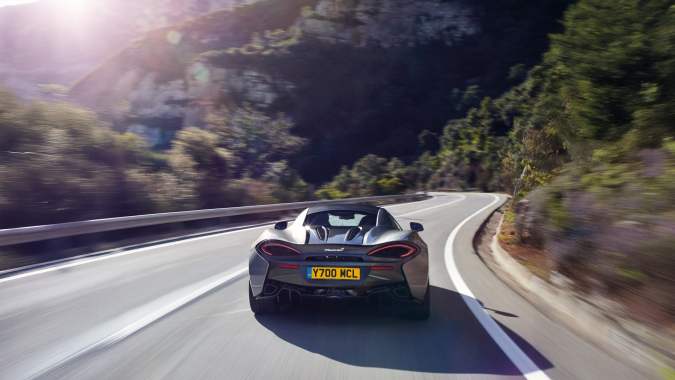 McLaren 570S gris circulando a gran velocidad por carretera de montaña con curvas, vista trasera dinámica al atardecer.