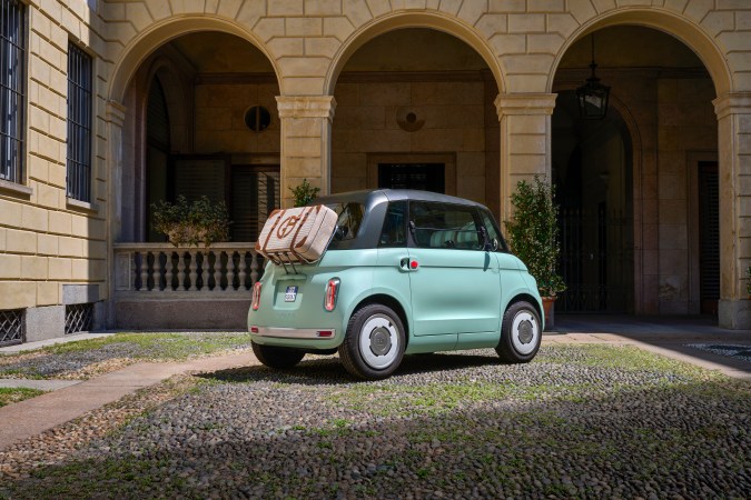 Fiat Topolino Dolcevita eléctrico con equipaje trasero, microcar urbano chic estacionado en edificio histórico italiano, estilo veraniego.