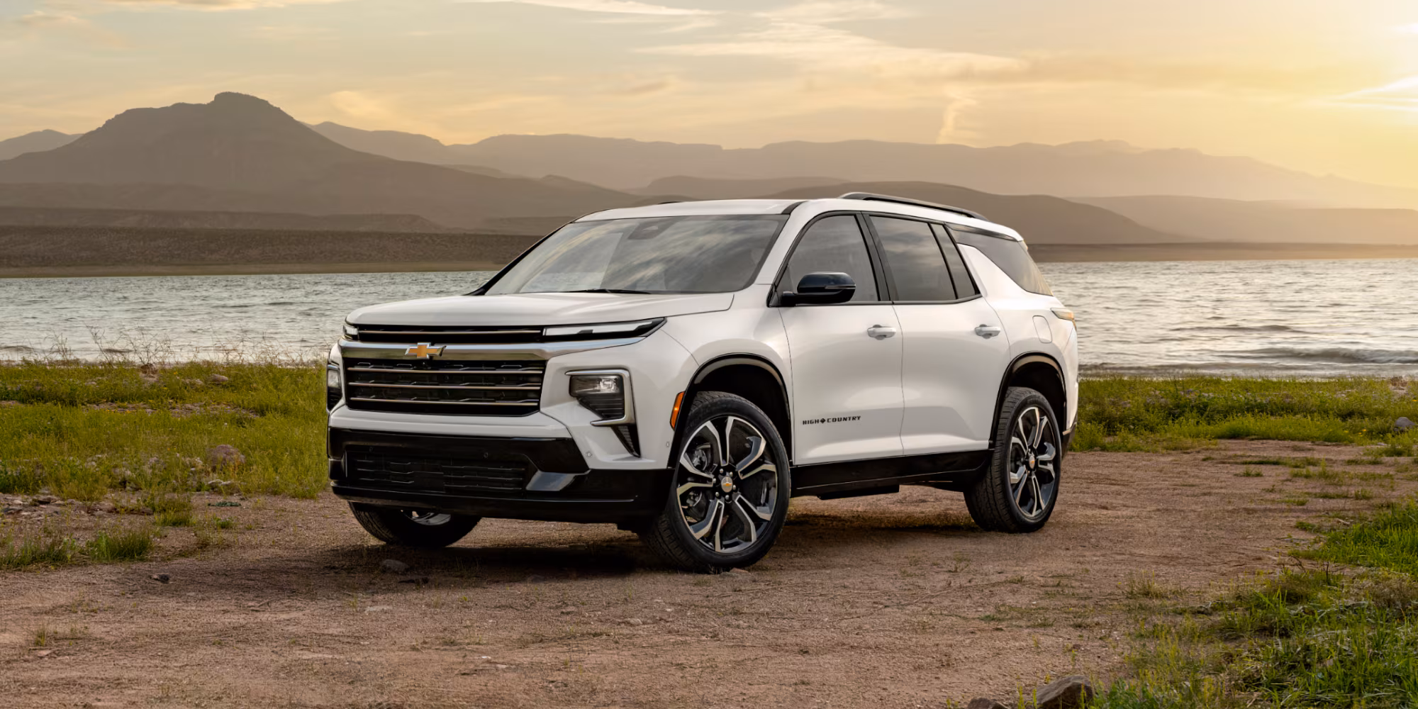Vista frontal del 2027 Chevrolet Traverse High Country blanco junto a un lago al atardecer.