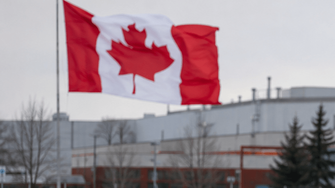 Autos estacionados frente a una planta industrial, con las banderas de Canadá y Estados Unidos ondeando y nieve alrededor.
