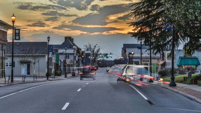 Avenida comercial con cámaras de seguridad, tráfico vehicular y comercios durante el atardecer.