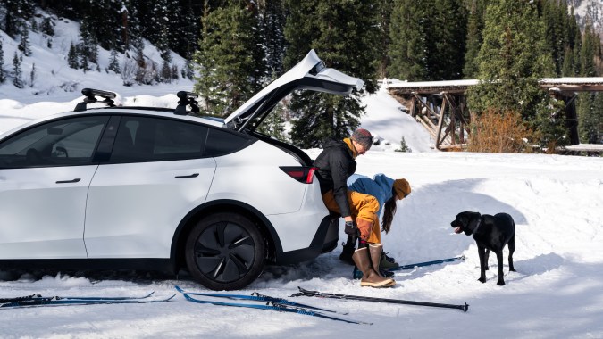 Tesla Model Y blanco con portón trasero abierto en un paisaje nevado, junto a una pareja, un perro y esquís.
