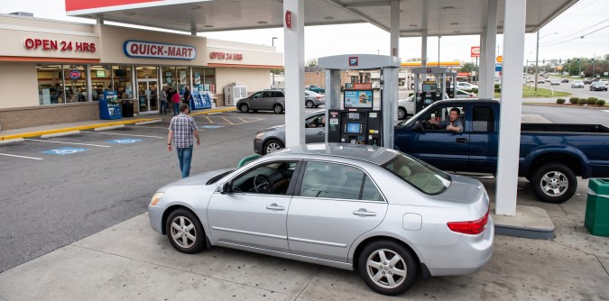 Automóviles y camionetas en gasolinera estadounidense con tienda Quick-Mart y surtidores de combustible.