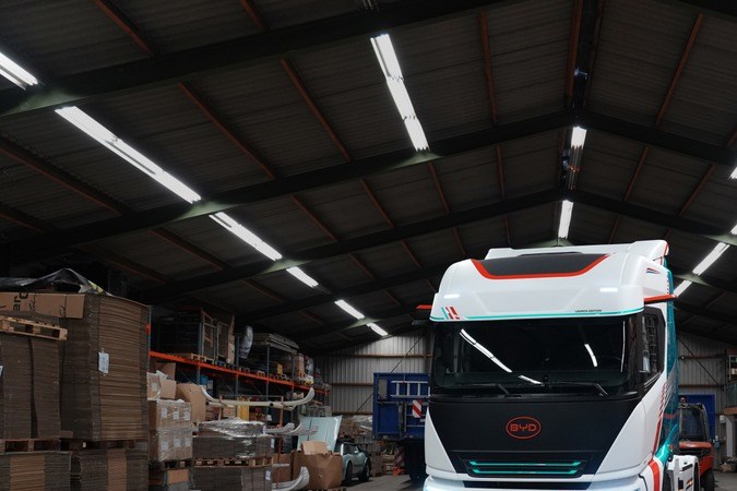 Camión eléctrico BYD ETT 44 con luces encendidas dentro de almacén logístico con estanterías de mercancía.