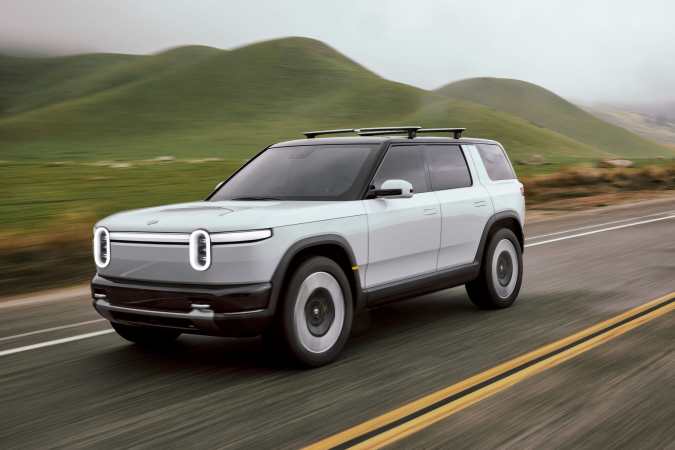 Rivian R2 blanco 2027 circulando por carretera con colinas verdes de fondo.