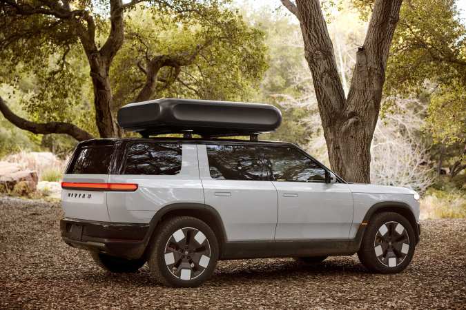 Rivian R2 blanco 2027 con cofre de techo estacionado entre árboles.