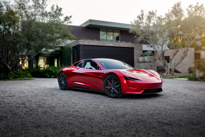 Tesla Roadster rojo estacionado frente a una casa moderna, con diseño deportivo, techo panorámico y detalles aerodinámicos.