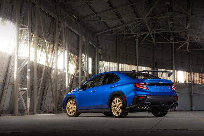 Subaru WRX tS azul con alerón, rines dorados y escape cuádruple en un almacén industrial.