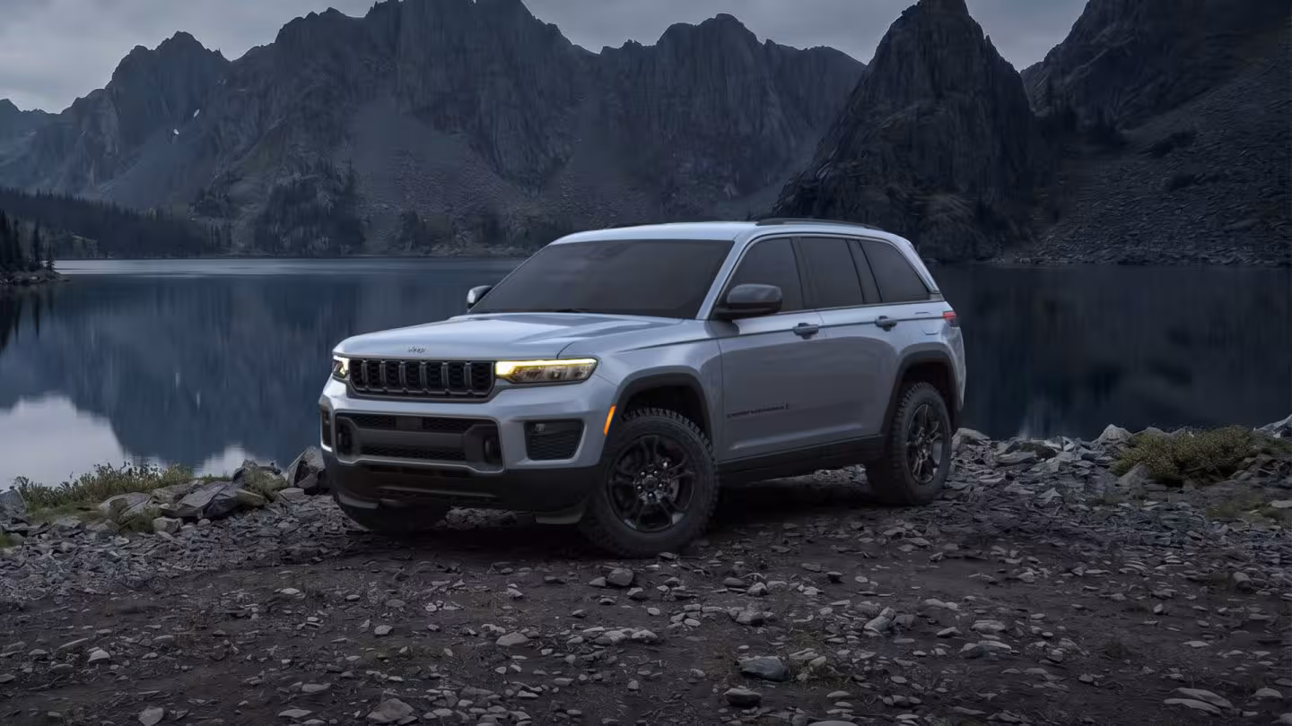 Camioneta SUV Jeep Grand Cherokee plateada en un paisaje montañoso junto a un lago alpino rocoso.
