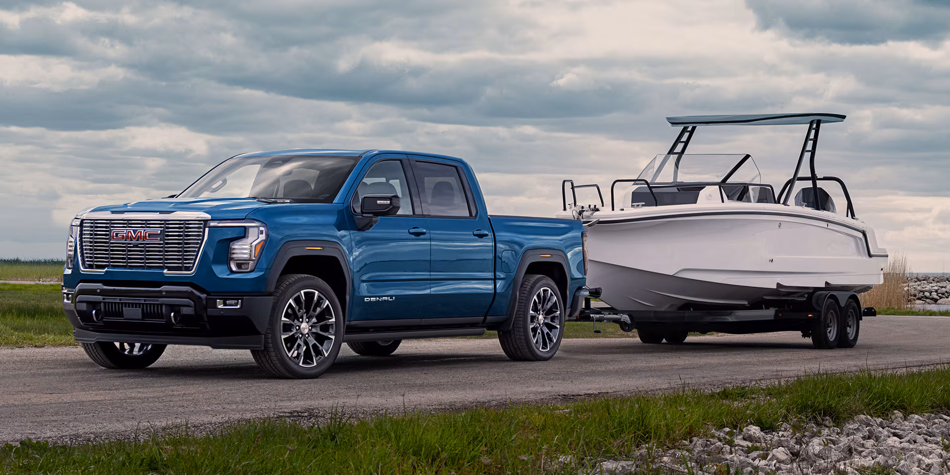 Camioneta GMC Sierra 1500 Denali azul remolcando un bote blanco sobre un tráiler en una carretera cerca del agua