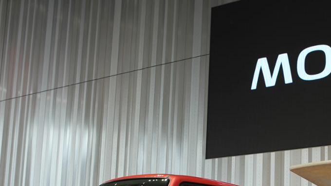 Presentación del Nissan Moco rojo en un evento automotriz, con un ejecutivo junto al vehículo.