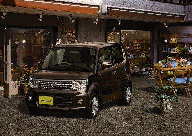 Nissan Moco negro estacionado frente a una cafetería, vehículo compacto japonés con diseño urbano.
