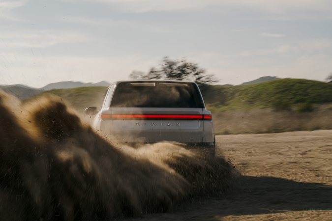 Rivian R2 blanco 2027 levantando polvo en camino de tierra, vista trasera.