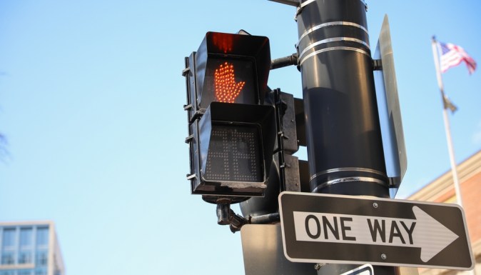 Semáforo peatonal rojo con mano iluminada, señal One Way y bandera estadounidense en ciudad.