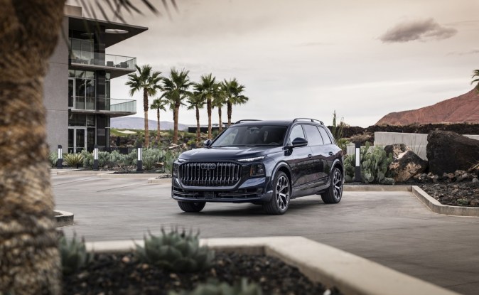 Audi Q9 2027 azul oscuro estacionado frente a un edificio moderno con palmeras, vista frontal de tres cuartos.