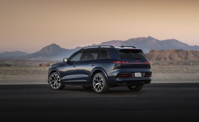SUV Audi Q9 azul oscuro visto de lado y desde atrás en una carretera desértica con montañas al fondo.