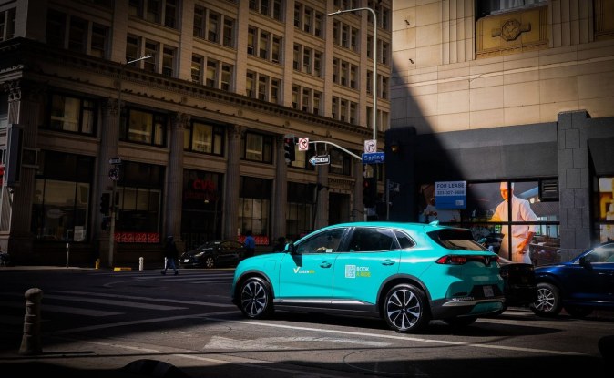 SUV VinFast VF8 turquesa con logotipos de Green SM circulando por una calle del centro de Los Ángeles.