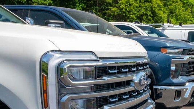 Fila de pickups pesadas Ford, Chevrolet y Ram junto a vans de carga blancas estacionadas en una pista de pruebas.