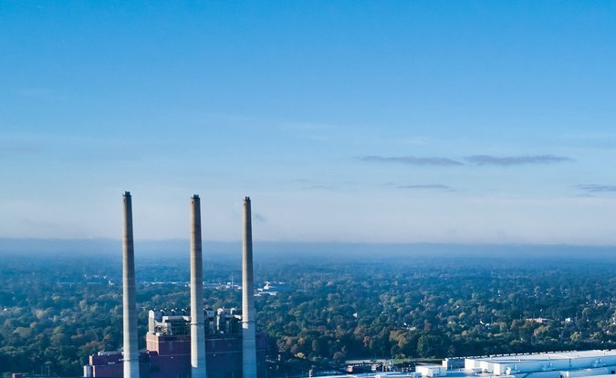 Vista aérea de un gran complejo industrial blanco rodeado de árboles, con tres chimeneas al fondo.