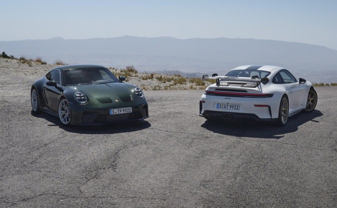 Dos Porsche 911 GT3, verde Touring y blanco con alerón, estacionados frente a montañas.