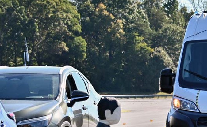 Van blanca Ram ProMaster avanza hacia un maniquí infantil que cruza la pista junto a autos estacionados durante una prueba.
