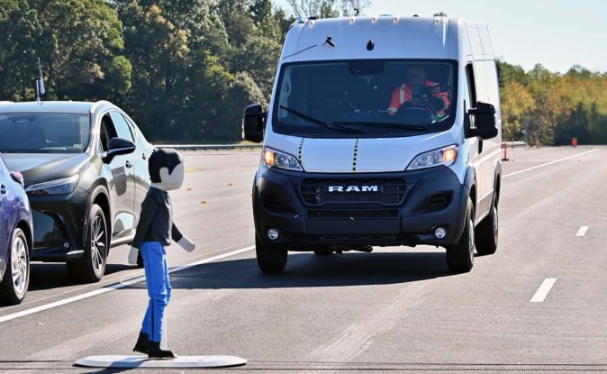 Van blanca Ram ProMaster avanza hacia un maniquí infantil que cruza la pista junto a autos estacionados durante una prueba.
