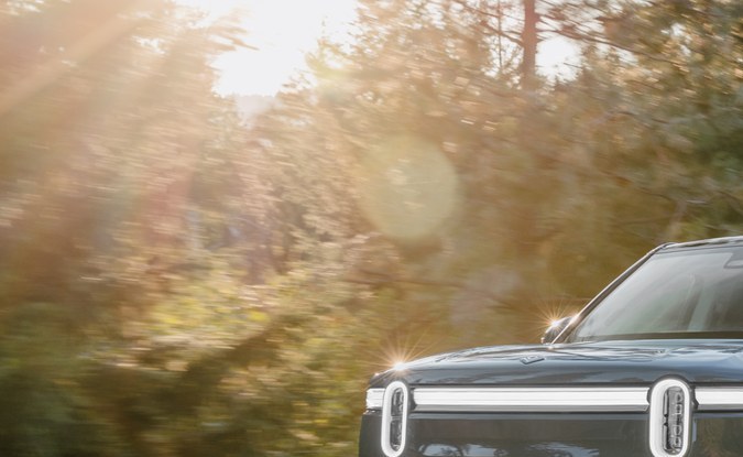 SUV eléctrico Rivian R1S gris circulando por una carretera rodeada de pinos con el sol de frente.