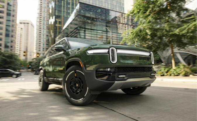 SUV Rivian R1S verde oscuro en una calle urbana rodeada de edificios altos.
