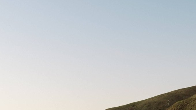 SUV eléctrico Rivian R2 azul circulando por una carretera de montaña con curvas al atardecer.