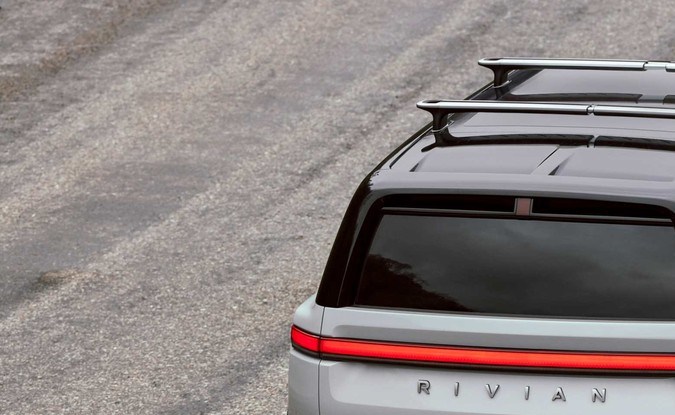 Vista trasera elevada de una Rivian R2 blanca con techo negro y barras portaequipajes en marcha.