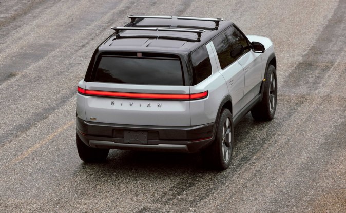 Vista trasera elevada de una Rivian R2 blanca con techo negro y barras portaequipajes en marcha.